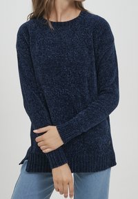 Femme portant un pull en tricot chenille bleu foncé avec des poignets et une bordure côtelés, associé à un jean bleu clair, les mains croisées à la taille.