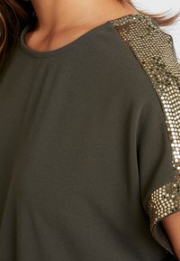 Ciemnozielona bluzka o gładkiej teksturze, z błyszczącymi złotymi cekinami na ramionach. Okrągły dekolt i krótkie rękawy.