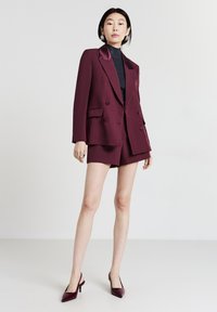 Blazer bordeaux doppiopetto con revers in raso, abbinato a pantaloni corti coordinati e décolleté a punta con cinturino. Top nero strutturato sottostante.