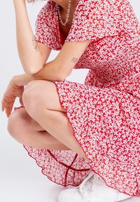 Robe florale rouge avec des motifs blancs, manches courtes, et un ourlet à volants. Assortie à des baskets blanches, mettant en valeur un tissu léger et une texture agréable.