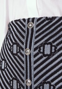 Jupe en tricot rayée noir et gris clair avec des boutons décoratifs argentés et des motifs floraux géométriques, associée à un chemisier blanc.