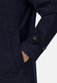 Mornarsko moder plašč z mehko teksturo, ki ima stransko žep in detajl z gumbom na manšeti, obložen z črnim gumbom.