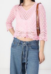 Top de punto rosa con escote en V, que presenta un patrón de encaje abierto y dobladillo con forma de ondas. Combinado con unos jeans de denim azul de cintura alta.