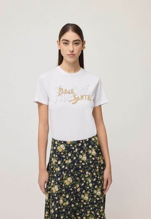 Frau mit langen dunklen Haaren trägt ein weißes T-Shirt mit gelbem Text und kleinen Sternen, kombiniert mit einem marineblauen Rock mit weißem Blumenmuster.