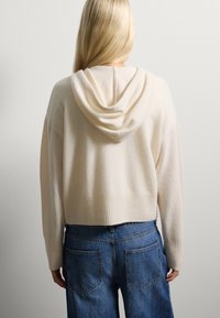 Felpa corta di colore crema con una consistenza morbida, dotata di cappuccio con coulisse e orlo a costine. Abbinata a jeans blu a vita alta.