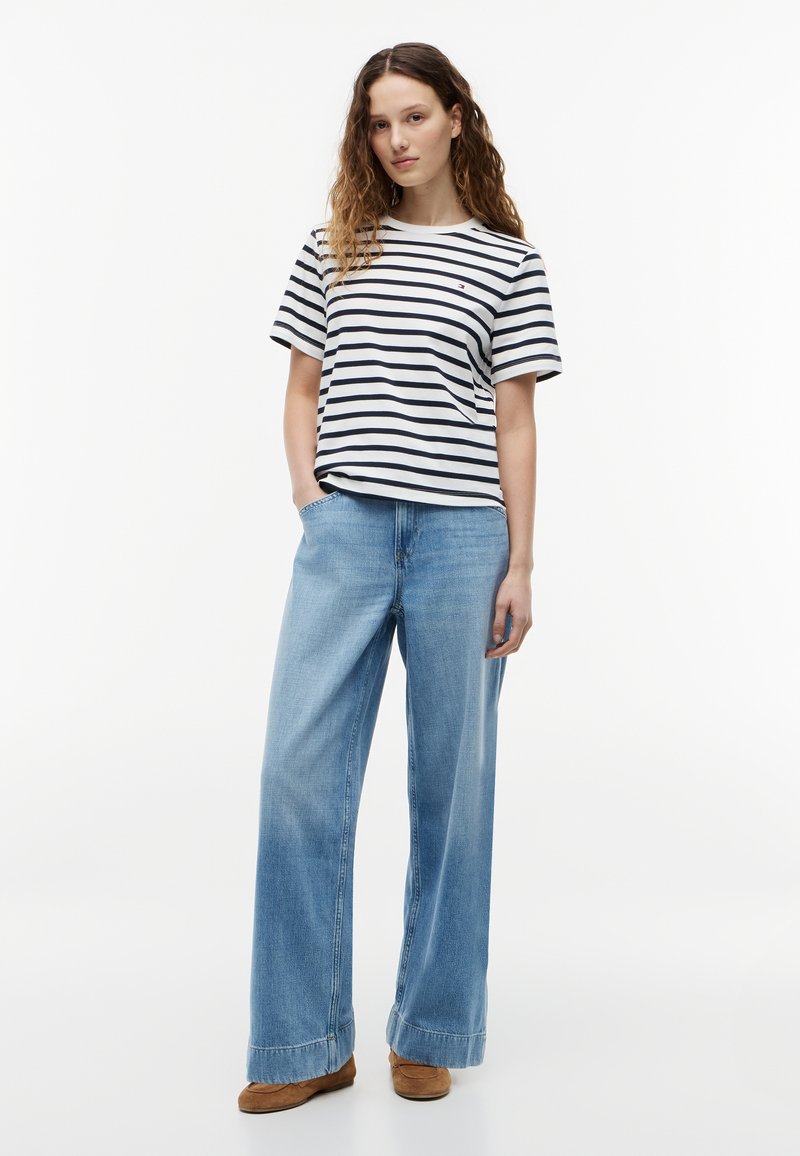 Femme aux longs cheveux bouclés portant un t-shirt à manches courtes rayé blanc et bleu marine, un jean large bleu clair et des chaussures marron, debout de manière détendue.