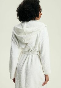 Biały, miękki szlafrok z teksturowanym wzorem, z kapturem, długimi rękawami i wiązaniem w pasie. Tkanina wydaje się być miękka i puszysta.