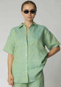Camicia verde chiaro a maniche corte con taglio rilassato, chiusura a bottoni sul davanti e un piccolo dettaglio del logo blu. Tessuto morbido, design casual.