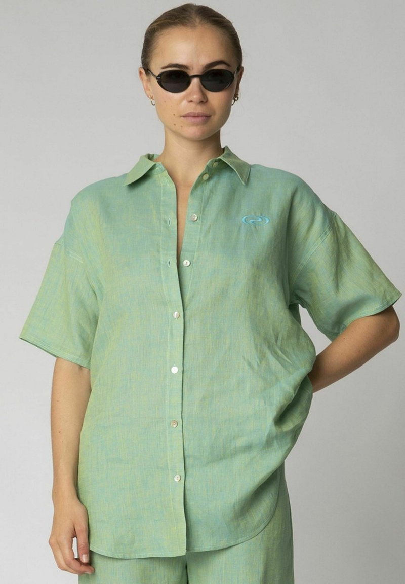 Camicia verde chiaro a maniche corte con taglio rilassato, chiusura a bottoni sul davanti e un piccolo dettaglio del logo blu. Tessuto morbido, design casual.