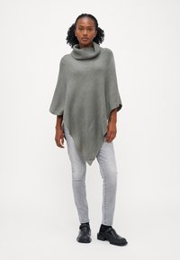 Grå strikket poncho med høy hals, med et teksturert mønster, asymmetrisk kant, brukt over lyse grå jeans og svarte sko.