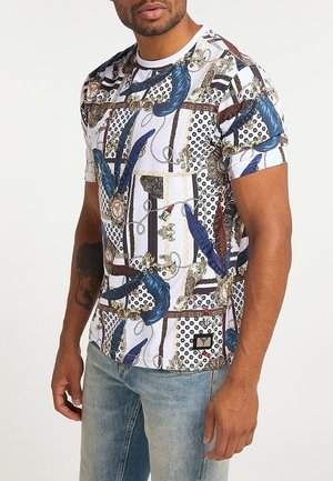 T-shirt blanc à manches courtes avec des motifs multicolores complexes, comprenant des plumes et des accents dorés. Présente un logo sur l'ourlet inférieur.