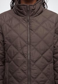 Gros plan d'une personne portant une veste matelassée marron avec un col montant et une fermeture éclair à l'avant.
