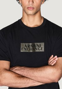Camiseta negra de cuello redondo con un logotipo rectangular dorado texturizado que dice "ARMANI EXCHANGE" en el pecho. Acabado de tela suave.