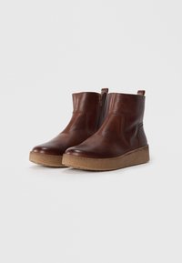 Botas de tobillo de cuero marrón con un acabado suave, cremallera lateral y puntera redonda, con una suela texturizada de un marrón claro y una asa en la parte posterior.