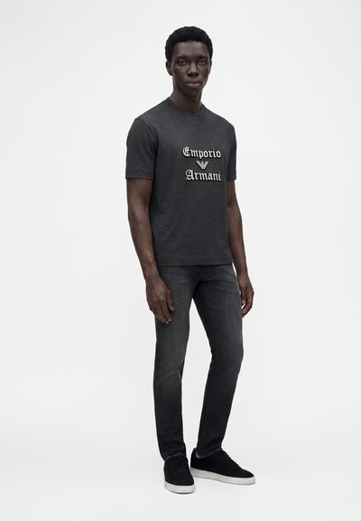 Νέος άνδρας που φοράει μαύρο t-shirt Emporio Armani, σκούρο γκρι τζιν και μαύρα αθλητικά με λευκές σόλες σε λευκό φόντο.
