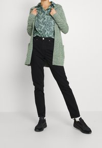 Světlý zelený dlouhý kardigan s kapsami, pod ním vzorovaná zelená košile, černé úzké džíny a černé tenisky z texturovaného materiálu.