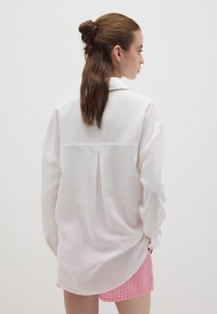 Camisa blanca de manga larga hecha de tela ligera, con un pliegue en la parte de atrás y un dobladillo curvo, combinada con una parte inferior de patrón rosa.