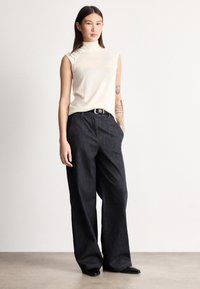 Ujjatlan törtfehér magasan záródó nyakú felső sötét farmer, széles szárú nadrággal párosítva, ezüst csattal ellátott övvel és elegáns fekete cipővel.