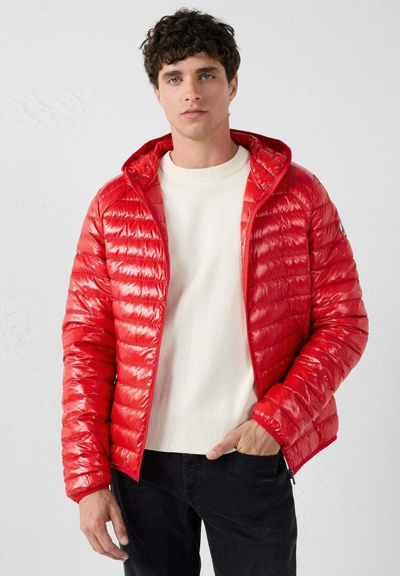 Giacca a piumino rossa con motivo trapuntato orizzontale, cappuccio e cerniera frontale. Abbinata a un maglione bianco girocollo e pantaloni neri.