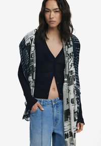 Schwarze gerippte Strickjacke mit Knöpfen vorne, kombiniert mit einem schwarz-weißen übergroßen Schal mit Textmustern und hellblauen Jeans.