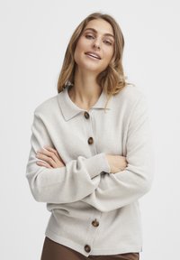 Ljusgrå cardigan med krage, prydd med bruna knappar, ribbade ärmslut och en mjuk strukturerad stickning. Bärs över en figurnära topp.