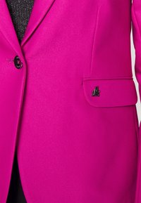 Fuchsiový blazer vyrobený z hladkého materiálu s jediným černým knoflíkem, levým kapsou a kovovým logem na kapse.