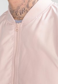 Närbild på en person som bär en ljusrosa zip-up-jacka, som visar en tatuerad nacke och en del av en grå skjorta under.