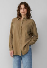 Khaki košile s knoflíky vyrobená z texturované látky, s dlouhými rukávy a volným střihem, mírně zastrčená do světle modrých džínů.