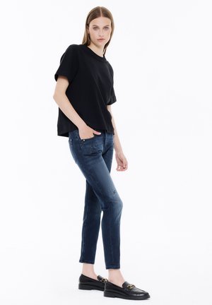 Schwarzes T-Shirt mit kurzen Ärmeln, dunkelblaue Jeans mit verwaschenen Akzenten und schwarze Slipper mit goldenen Details. Schlichtes, modernes Design.