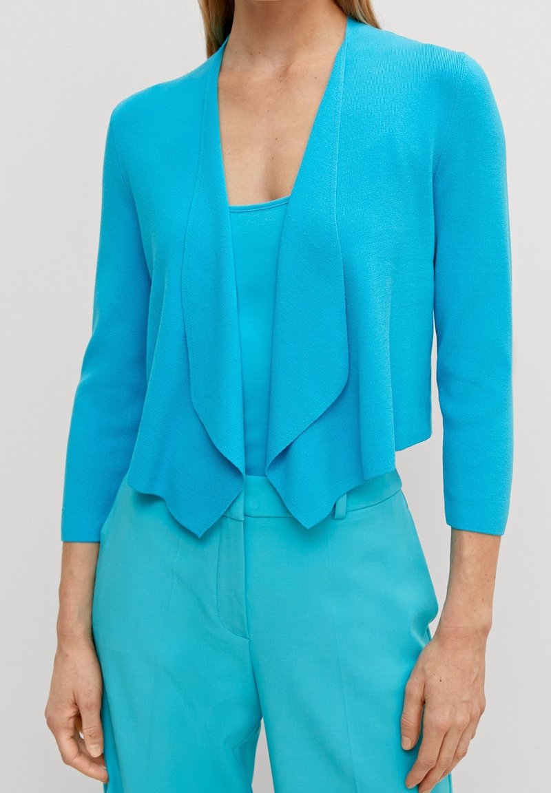 Cardigan turquoise ouvert avec un design drapé, texture douce et manches trois-quarts, porté sur un haut et un pantalon assortis.