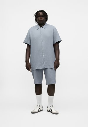 Homme avec dreadlocks portant une chemise à manches courtes bleu clair boutonnée et un short assorti, des chaussettes blanches et des baskets blanches avec des bandes noires.