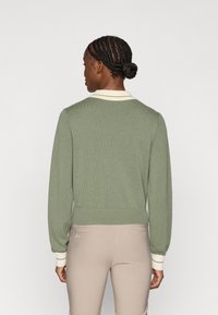 Zaļš adīts džemperis ar krēmkrāsas apkakli un aproču akcentiem, ar pieguļošu jostu un teksturētu virsmu. Skats no aizmugures.