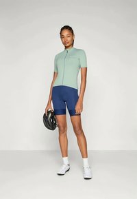 Completo da ciclismo composto da una maglia verde chiaro a maniche corte con zipper frontale e pantaloni corti blu navy. Gli accessori includono un casco nero e scarpe bianche.