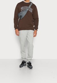 Sudadera marrón con texto "WOODBIRD", bolso bandolera gris, pantalones deportivos grises y zapatillas negras Nike. Conjunto casual y cómodo.