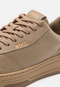 Beige nahka sneaker, jossa on teksturoitu pinta, hillitty ompelu ja vastaavat nauhat. Varustettu kohotetulla logolla ja kumipohjalla.