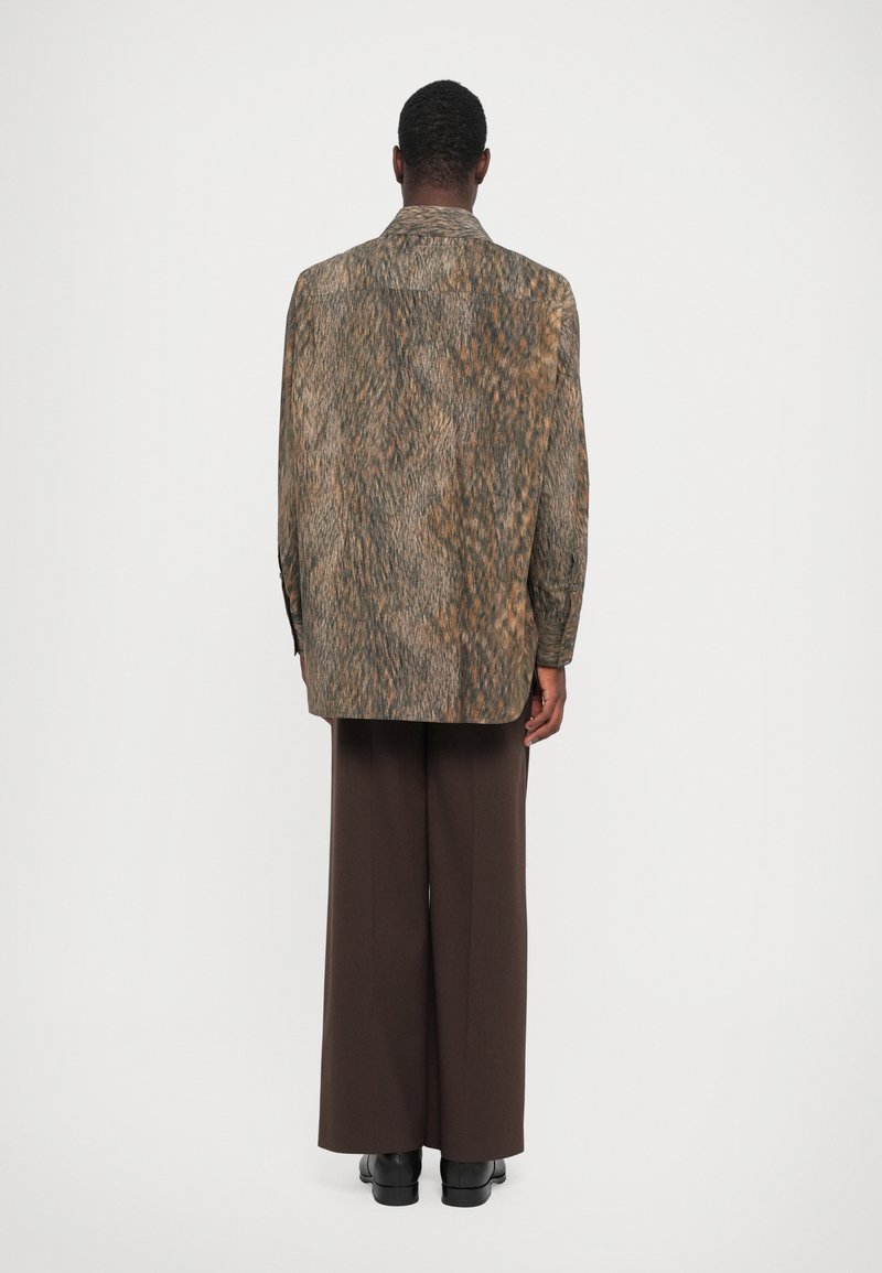 Persoon die met de rug naar de camera staat, gekleed in een gestreepte lange mouw shirt, brede bruinbroek en zwarte schoenen, tegen een effen achtergrond.