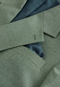 Primo piano di una giacca verde testurizzata con rever a incavo, tasca e fodera interna blu scuro con motivo visibile.