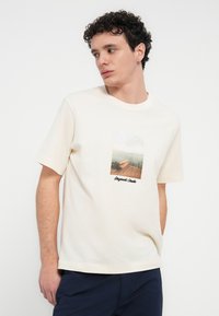 Μπεζ βαμβακερό t-shirt με κοντά μανίκια, με μπροστινό γραφικό που απεικονίζει μια θαλάσσια σκηνή και το κείμενο "Originals Studio" από κάτω.