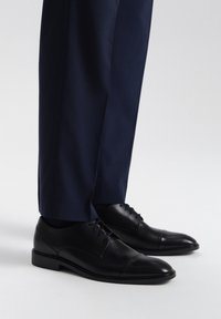 Chaussures habillées en cuir noir avec une finition lisse et un bout en cap, associées à un pantalon texturé bleu marine, mettant en avant une tenue formelle.
