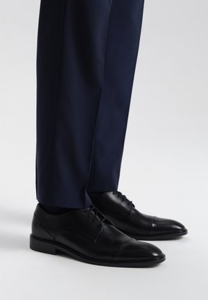 Chaussures habillées en cuir noir avec une finition lisse et un bout en cap, associées à un pantalon texturé bleu marine, mettant en avant une tenue formelle.
