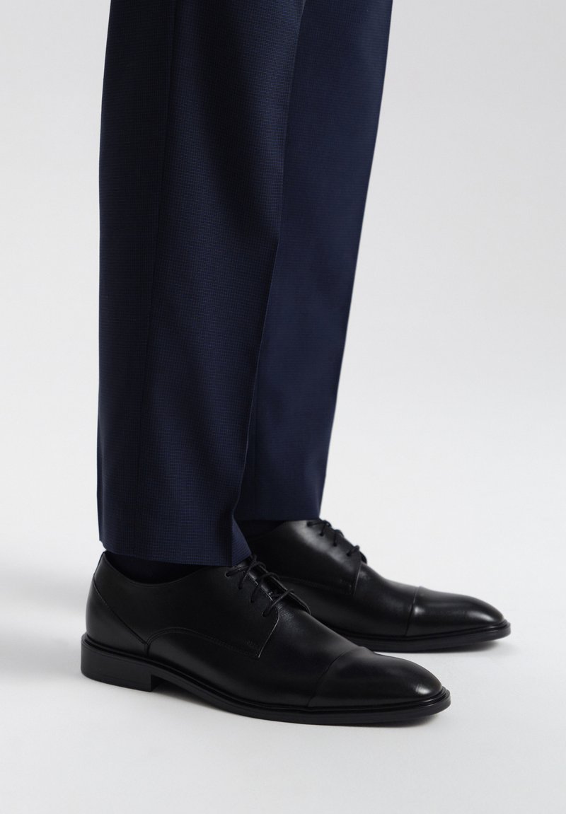 Chaussures habillées en cuir noir avec une finition lisse et un bout en cap, associées à un pantalon texturé bleu marine, mettant en avant une tenue formelle.
