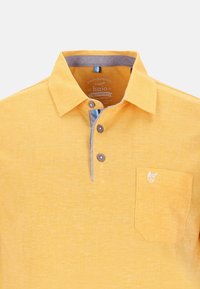 Polo shirt jaune en tissu texturé, avec un col gris, trois boutons marron et un petit logo blanc sur la poche gauche.