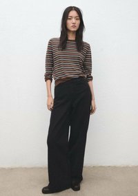 Femeie în picioare în fața unui perete deschis la culoare, purtând o cămașă cu dungi maro cu mâneci lungi, pantaloni negri cu picioare largi și pantofi negri.