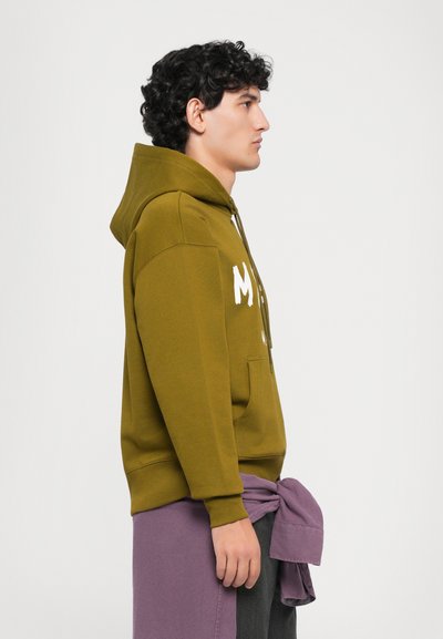 Jaunas vyras olivinės spalvos hoodie su baltu tekstu, purpurinė džemperis surištas aplink liemenį, atsigręžęs į dešinę priešais baltą fono.