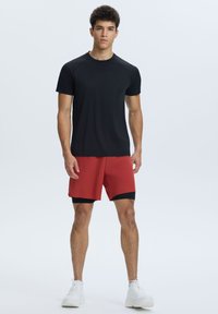Camiseta atlética negra de manga corta con mangas raglán, combinada con pantalones cortos rojos con un acento negro. Modelo con zapatillas blancas.