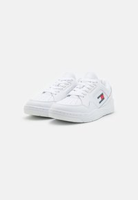 Zapatillas blancas de piel sintética con puntera perforada, suela gruesa y un detalle de logo en rojo, blanco y azul en el lateral.