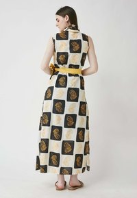 Maxi dress with a sleeveless design, featuring a bold leaf pattern in black and golden on a white background. Tied yellow belt accentuates waist.