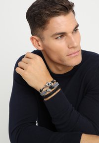 Sorte og grå multi-strengs armbånd med vævede strukturer, perler og metaldetaljer, båret på et håndled med en mørkfarget sweater.