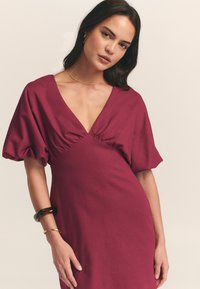 Vestido burdeos con un escote en V profundo, detalle de busto fruncido y mangas abullonadas. Textura suave con una silueta ajustada. Acento de pulsera dorada.