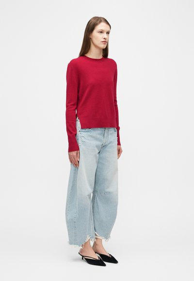 Γυναίκα που φοράει κόκκινο πουλόβερ με μακριά μανίκια, ανοιχτό μπλε φαρδιά τζιν παντελόνια με φθορές και μαύρα μυτερά πέδιλα με λουρί, στέκεται μπροστά σε λευκό φόντο.